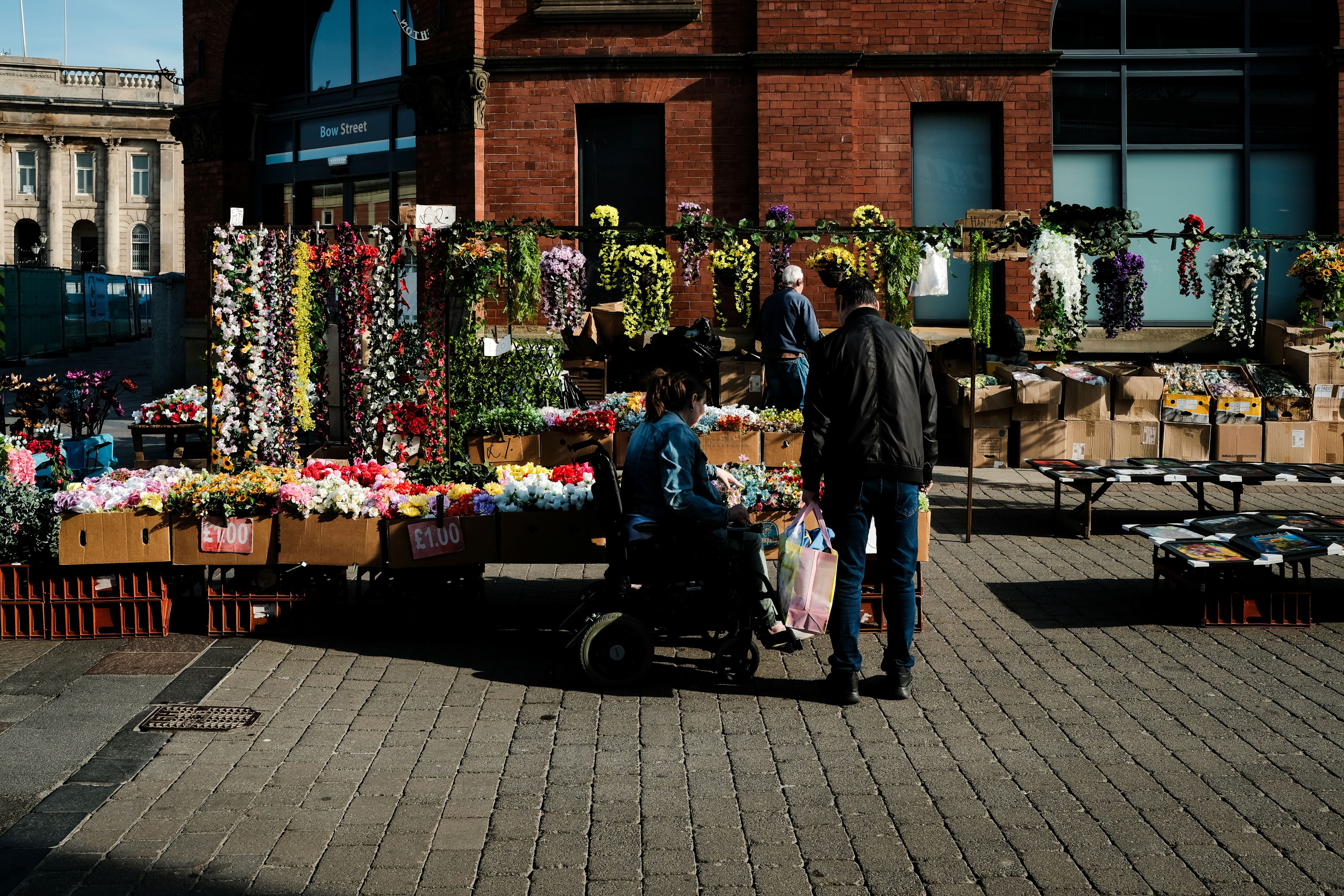 A woman who uses a wheelchair next to a man. They are viewing flowers that are set out on tables on the pavement as part of a market stall..