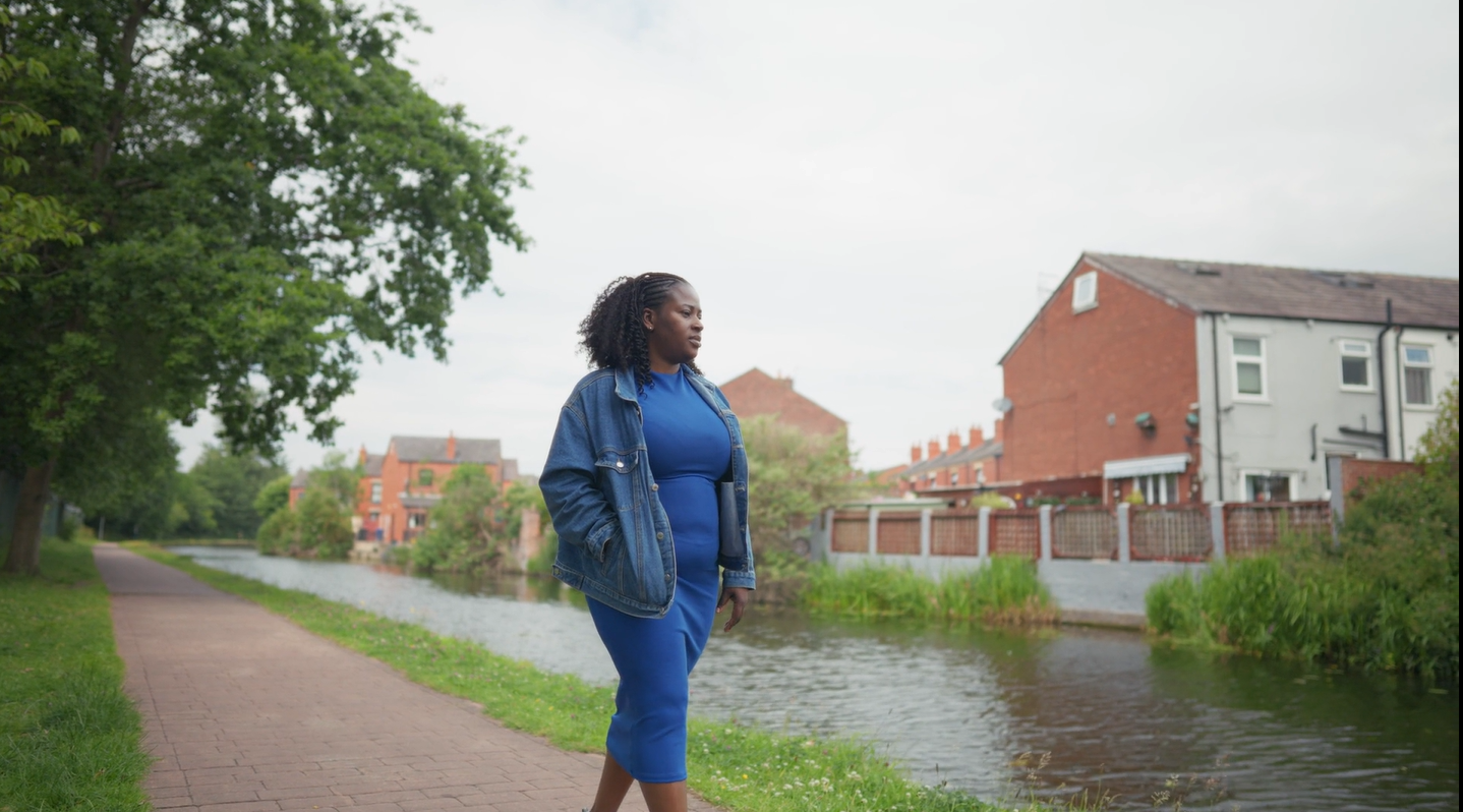 Person walking beside a canal lined with trees and red-brick homes in an urban setting.