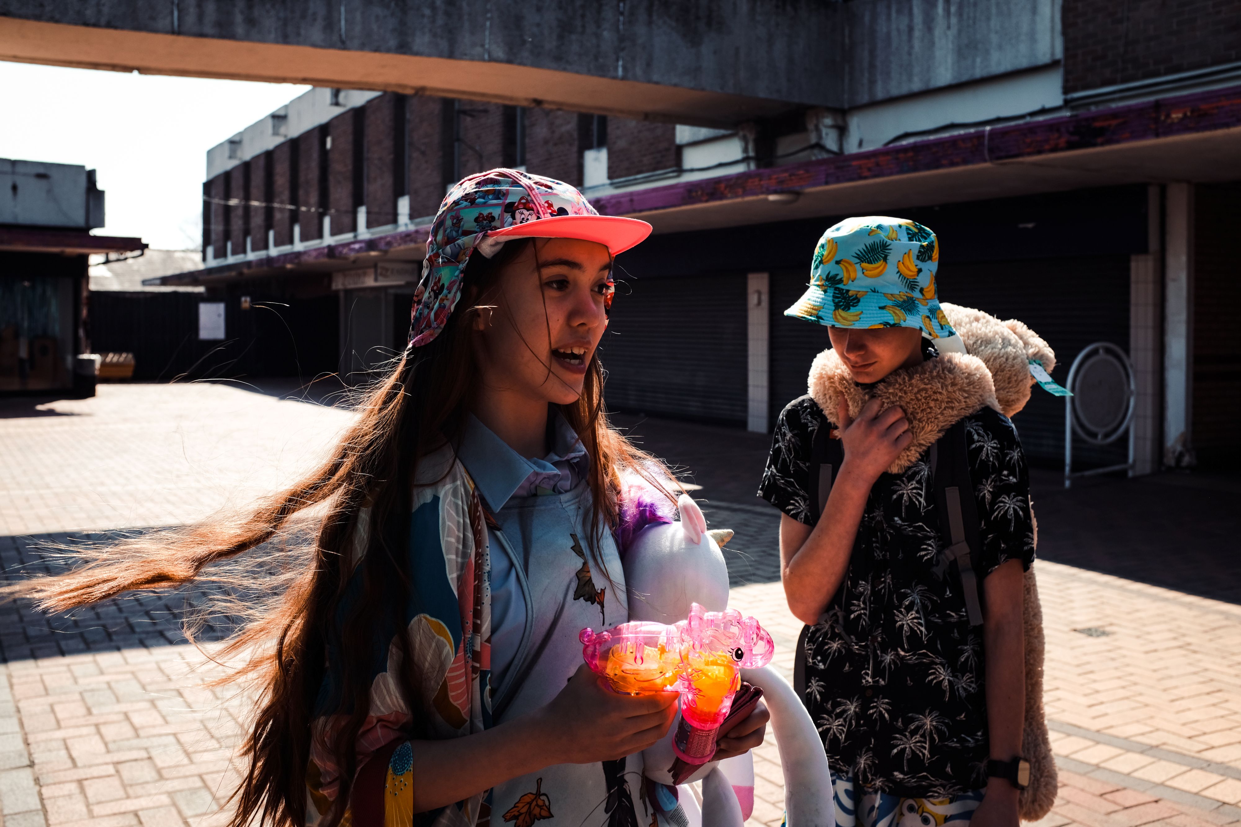 Two children in front of closed shop shutters; a girl in a floral cap holds an inflatable unicorn and a pink toy while a boy in a banana‑print bucket hat looks down with a teddy on his shoulders