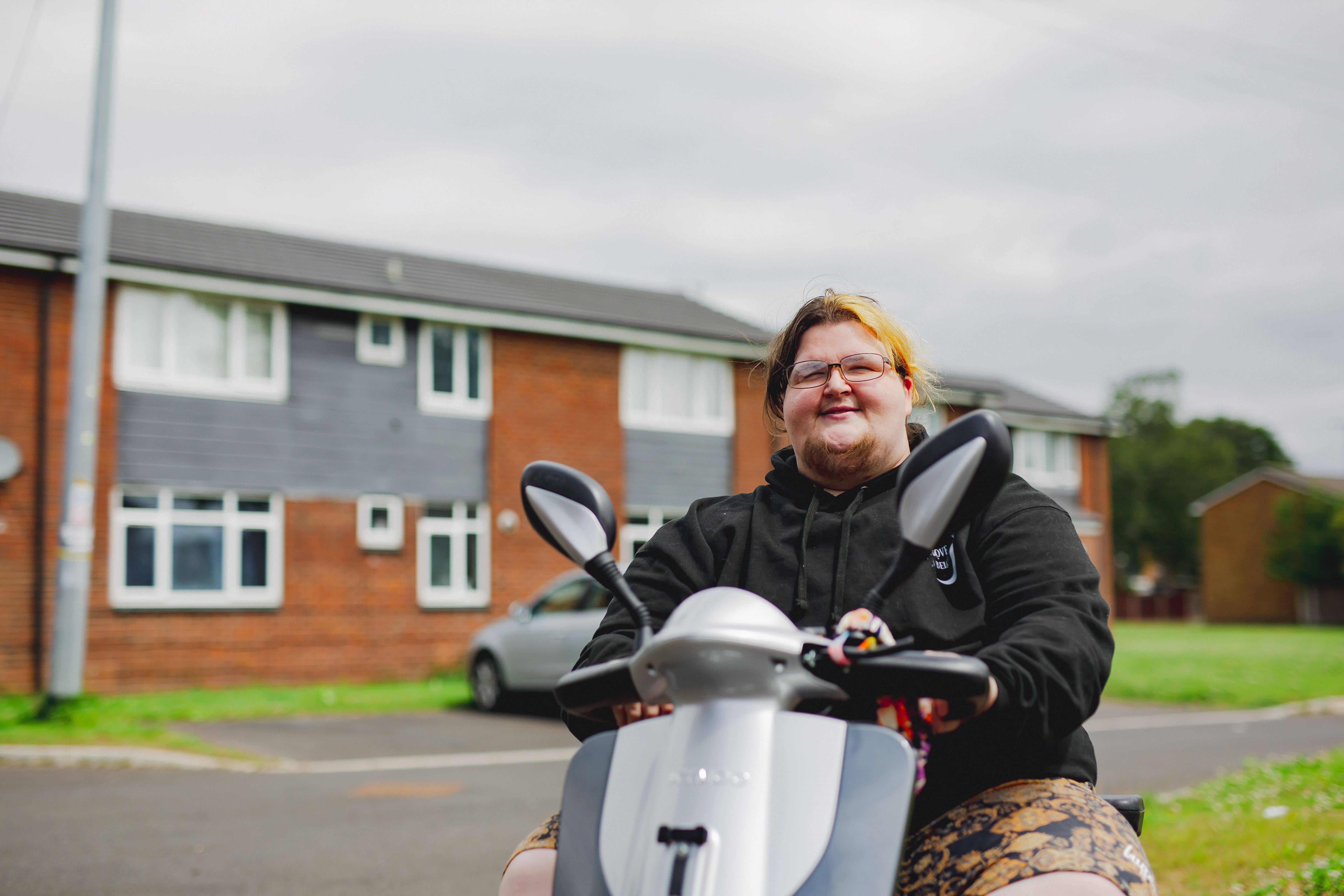 Person on a scooter in a suburban neighborhood, with brick houses and parked cars in the background.