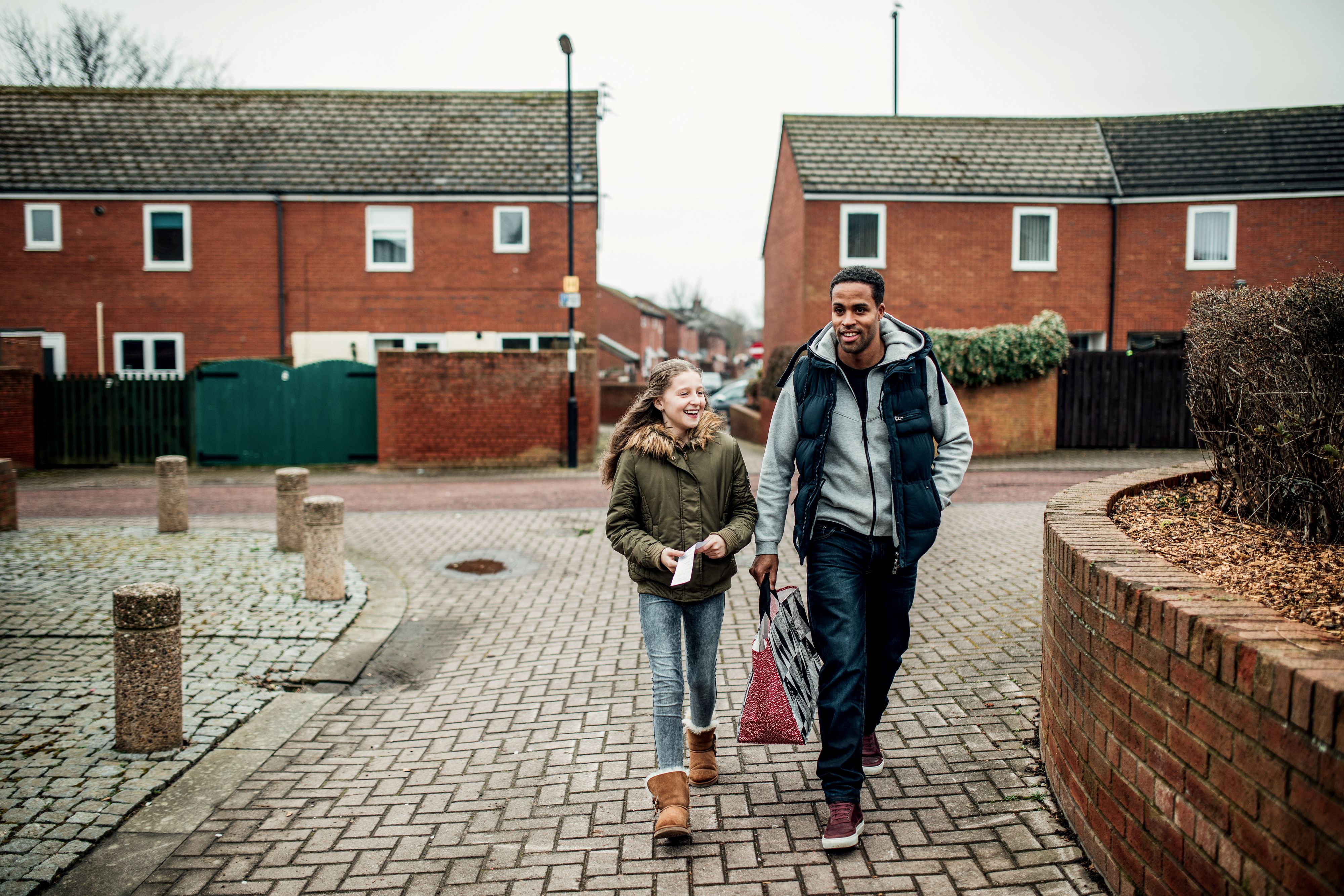 A smiling man and a young girl walk side by side along a street of red‑brick houses on an overcast day