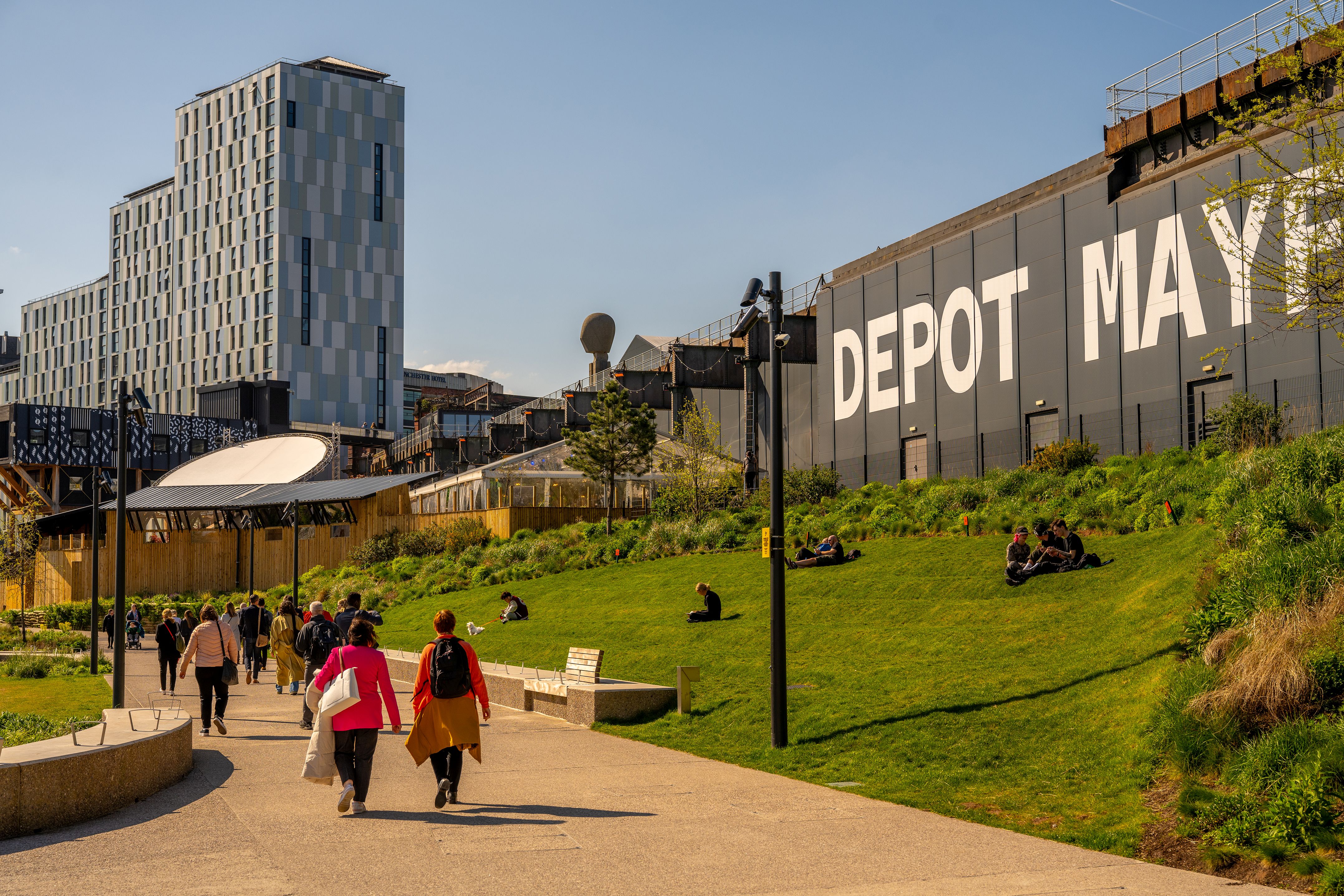 Urban green space Depot Mayfield in Manchester, people on a paved path and small groups sitting on a grassy slope beside a tall industrial wall.