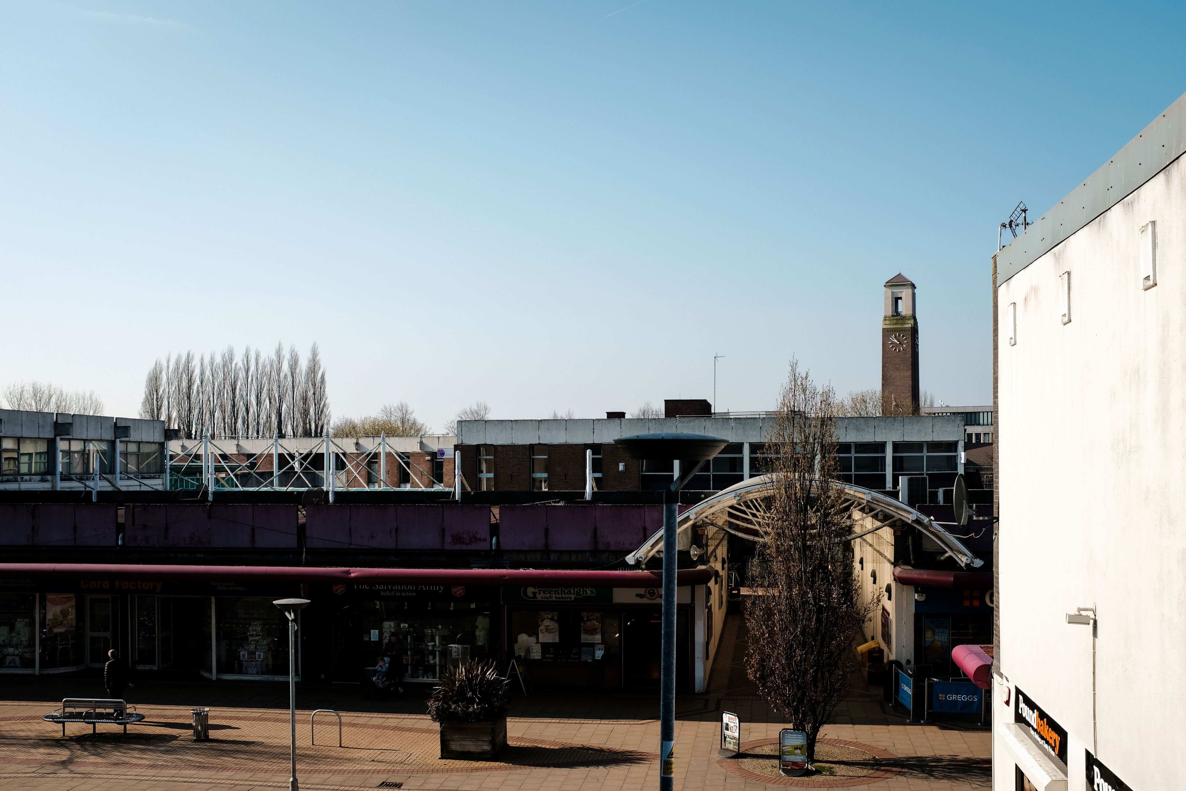 Salford shopping centre, a covered pedestrian arcade entrance, leafless trees and a tall brick clock tower against a clear blue sky