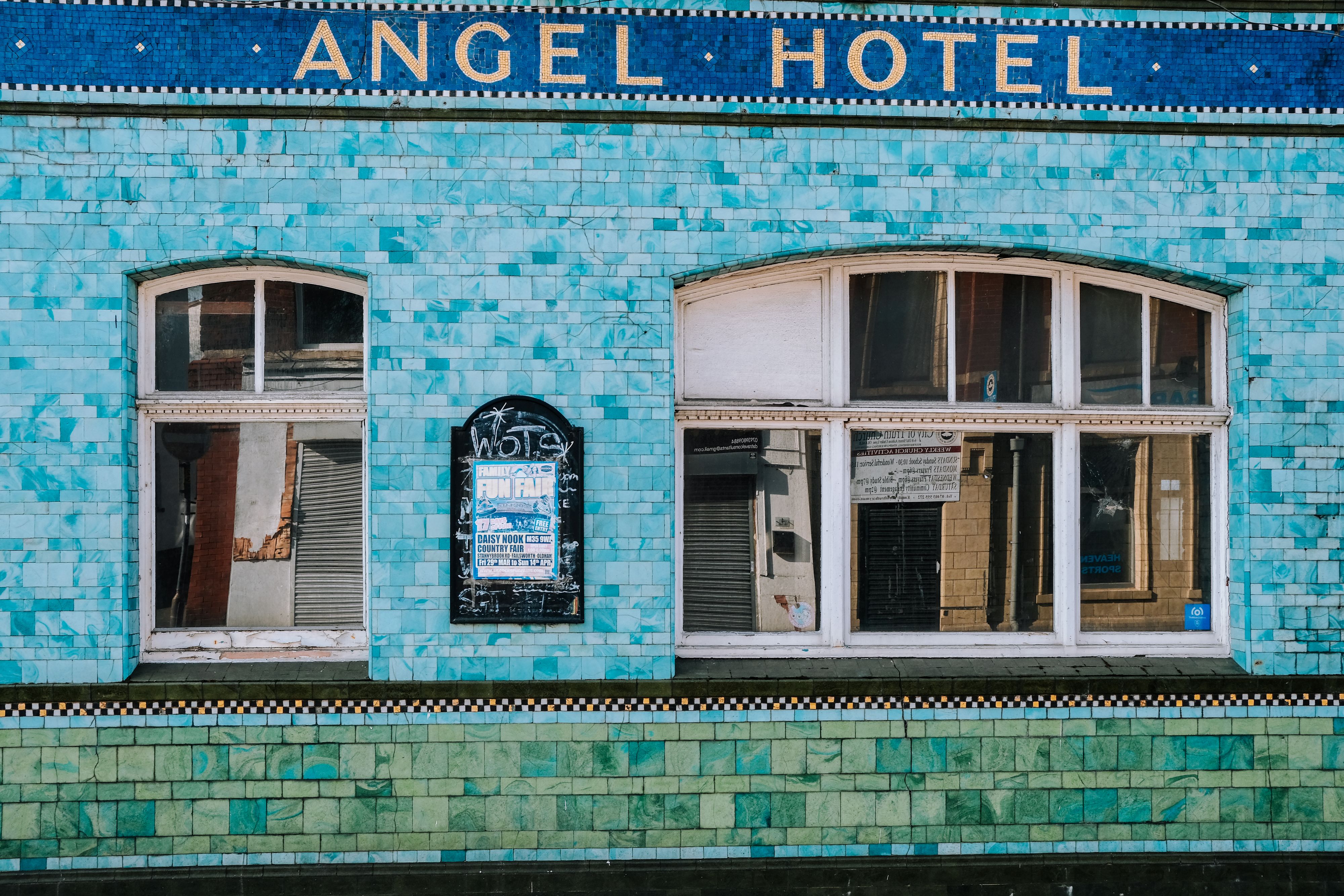 Facade of a tiled building covered in small turquoise and green ceramic tiles with a blue mosaic band reading "ANGEL HOTEL"